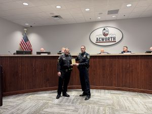 Two uniformed police officers stand in a meeting room, one presenting a plaque to the other. An “Acworth” sign and American flag are visible in the background.