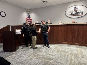 A man shakes hands with a police officer in a meeting room. Other officials are seated behind a wooden desk with the word "Acworth" on the wall.