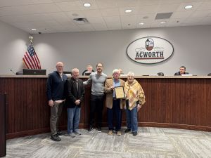 Five people standing in front of a council desk, with one person holding a framed document. An American flag and an "Acworth" signage are behind them.