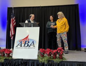 Three people stand on a stage at an Acworth Business Association event. The person on the left speaks at a podium, while the two on the right hold hands. A U.S. flag and poinsettias are visible.