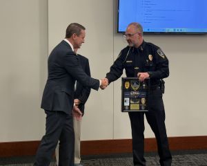 A man in a suit shakes hands with a uniformed officer holding a plaque in a conference room with a large screen behind them.