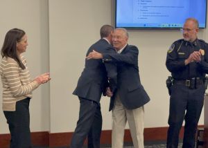 A man in a suit hugs another man while two people, a woman and a man in a police uniform, stand nearby watching in a room with a screen displaying text.