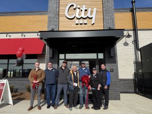 A group of seven people stand in front of a City BBQ restaurant, some holding red ribbon and scissors. Red and black balloons are on the left.