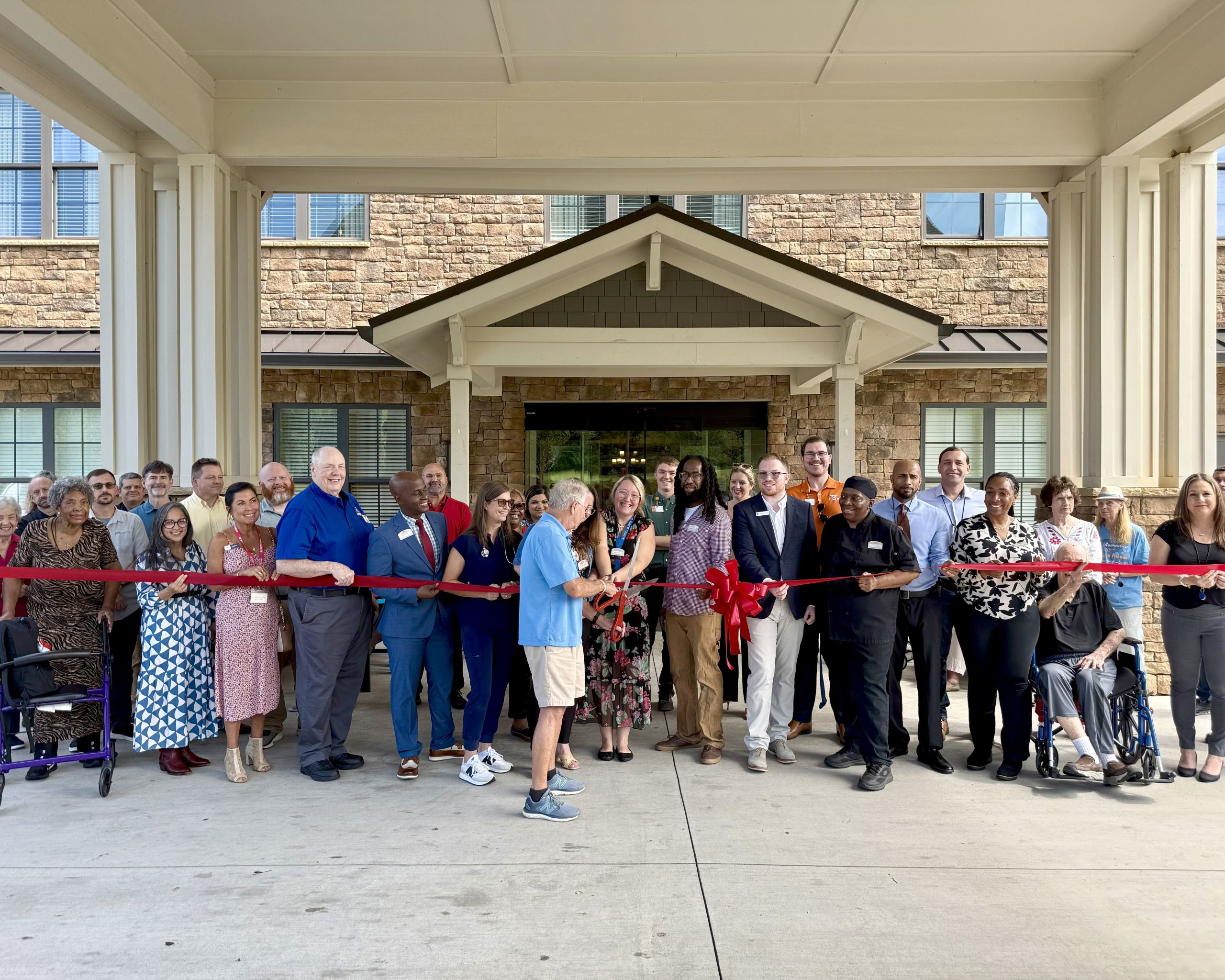 A large group of people stands in front of a building’s entrance, holding a red ribbon for a ribbon-cutting ceremony. Two people in the center are cutting the ribbon.