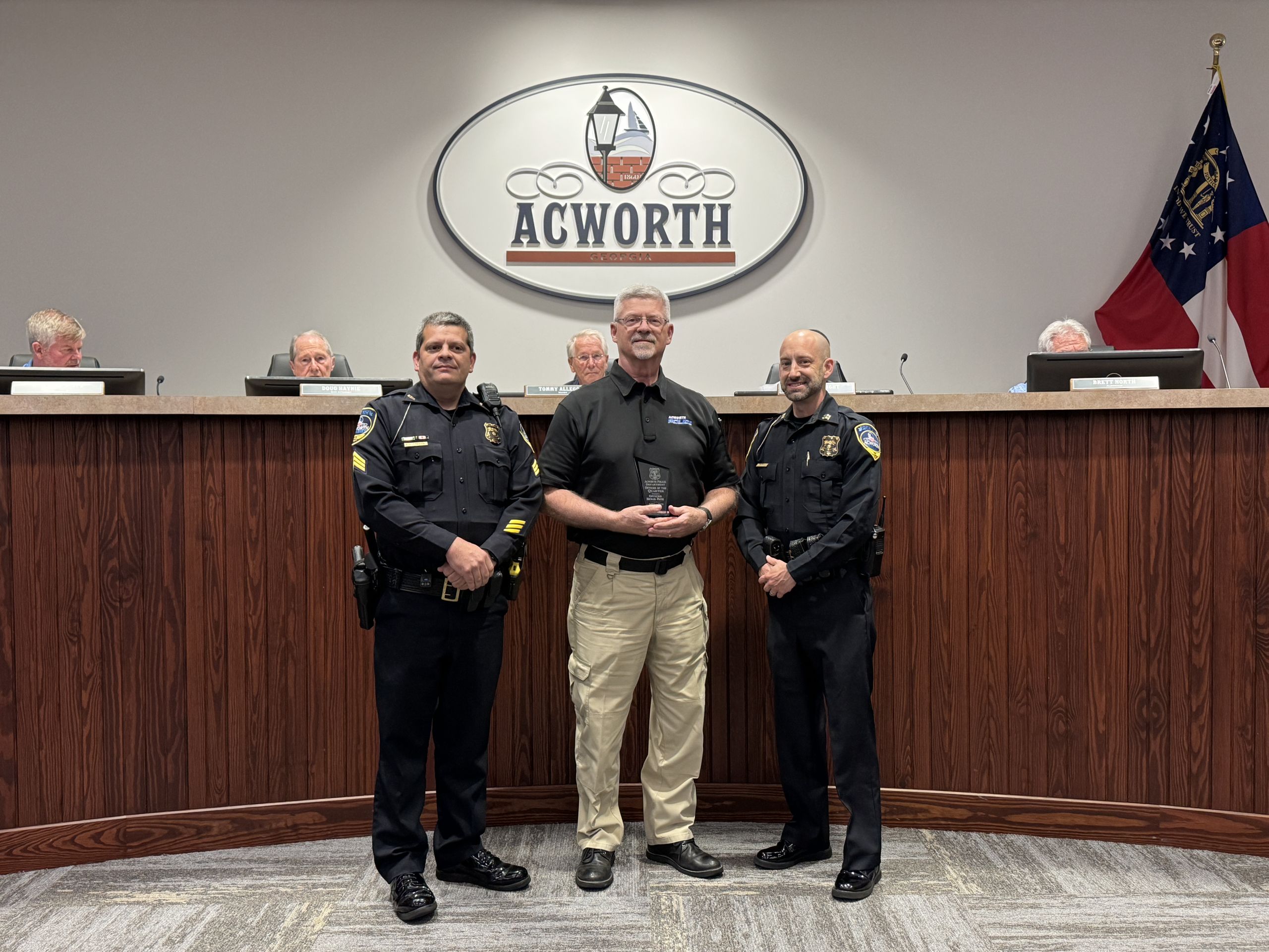Three men stand in front of a wooden podium with an "Acworth" sign; two are in police uniforms and one is in civilian clothing. Four people are seated at the podium in the background.