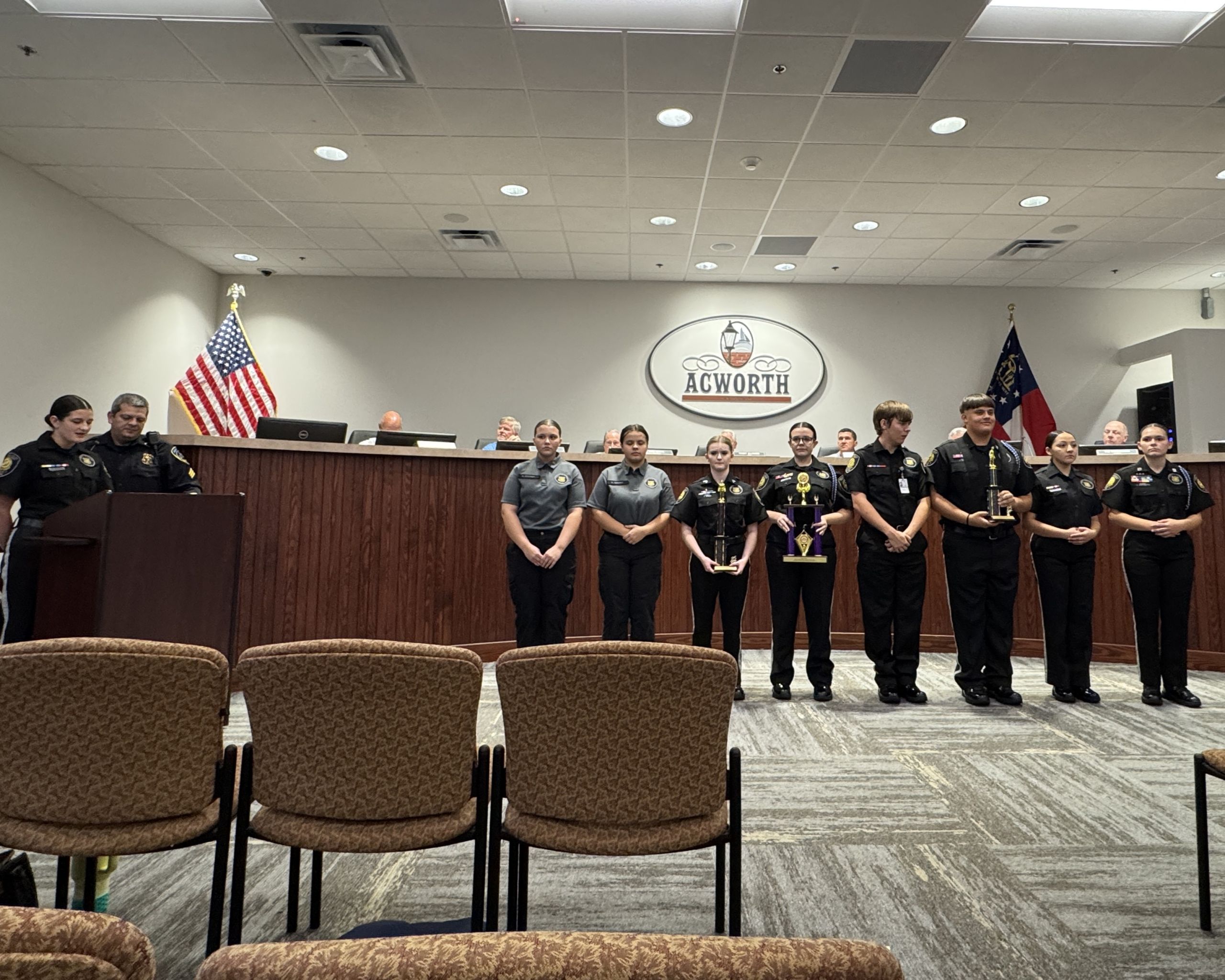 A group of uniformed officers stands in a row facing the audience in a government meeting room with Acworth signage and two U.S. flags on the wall behind them.