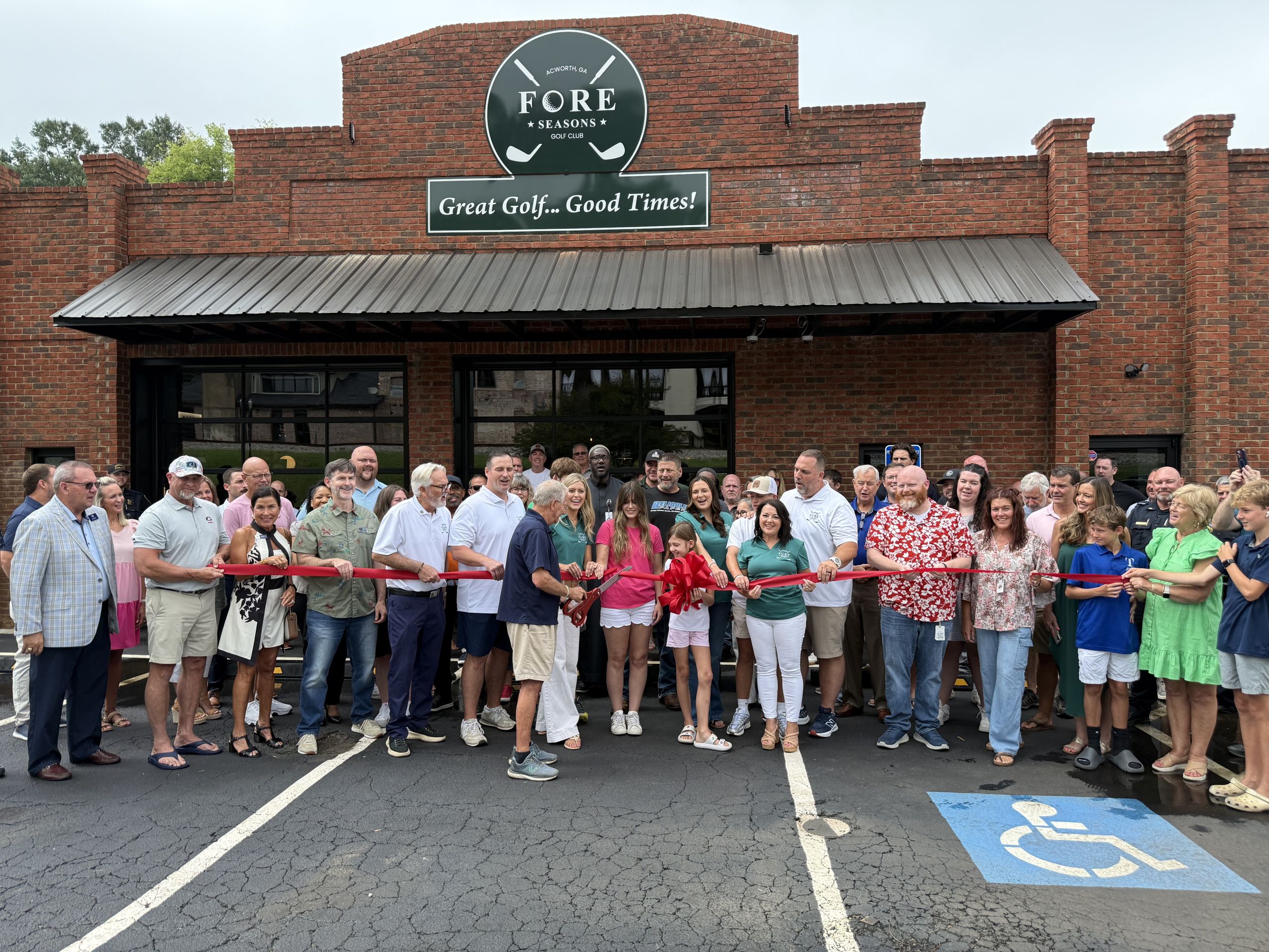A large group of people pose for a ribbon-cutting ceremony in front of a brick building with a sign that reads "FORE Seasons.