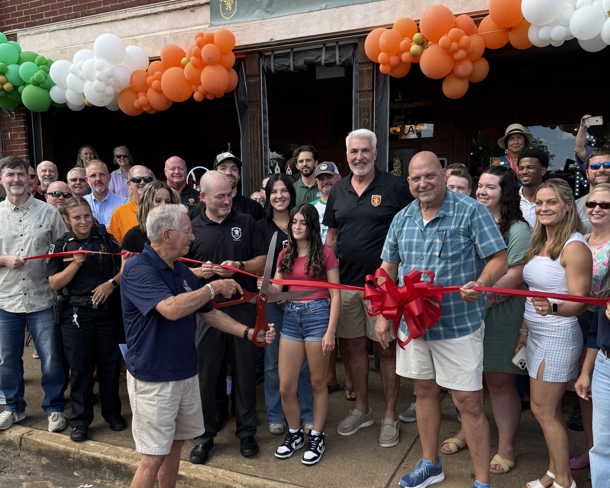 A group of people stand in front of a building decorated with balloons while a man cuts a large red ribbon for a ribbon-cutting ceremony.