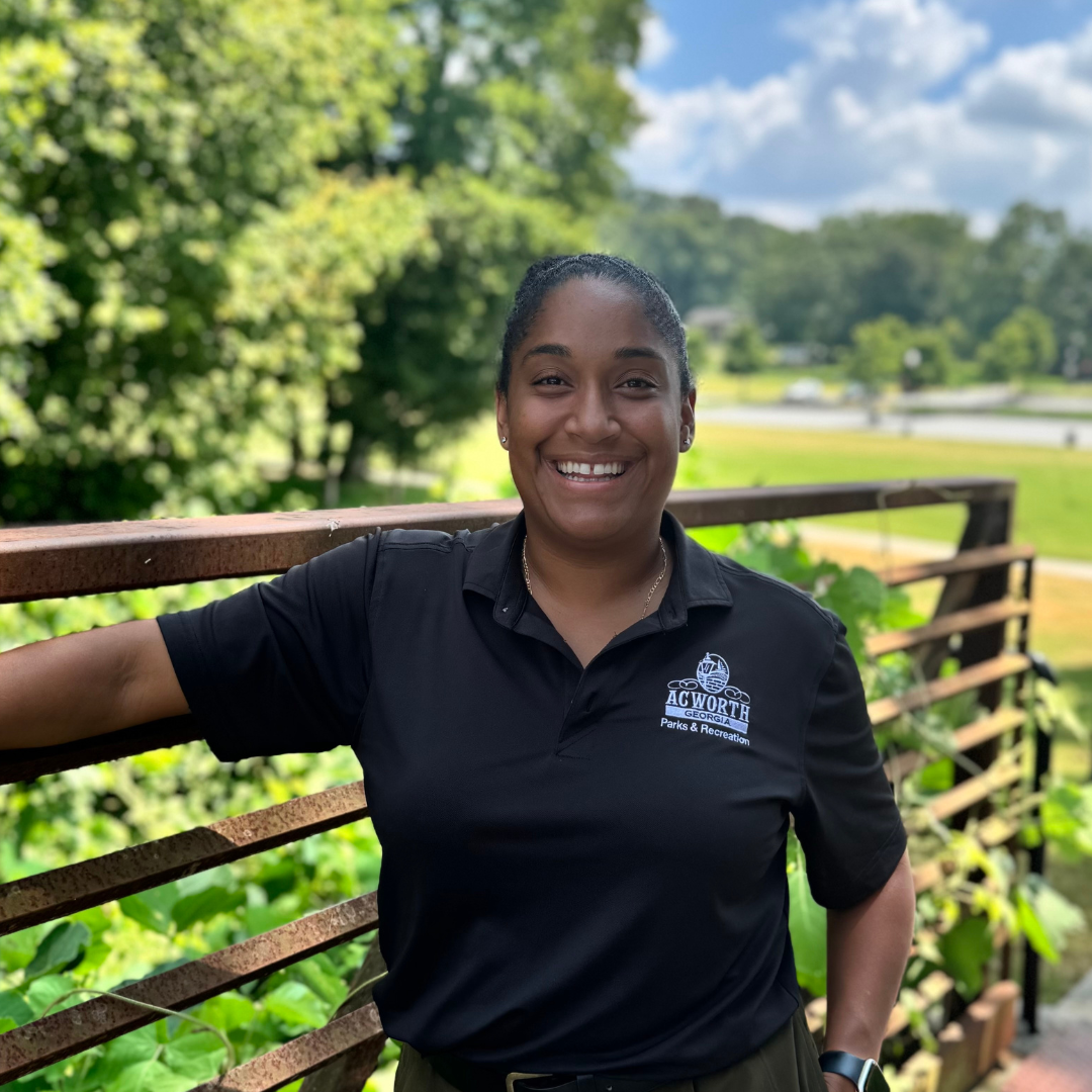 A person in a black "Acworth Parks & Recreation" polo stands outdoors by a metal railing, smiling, with greenery and a lake in the background.