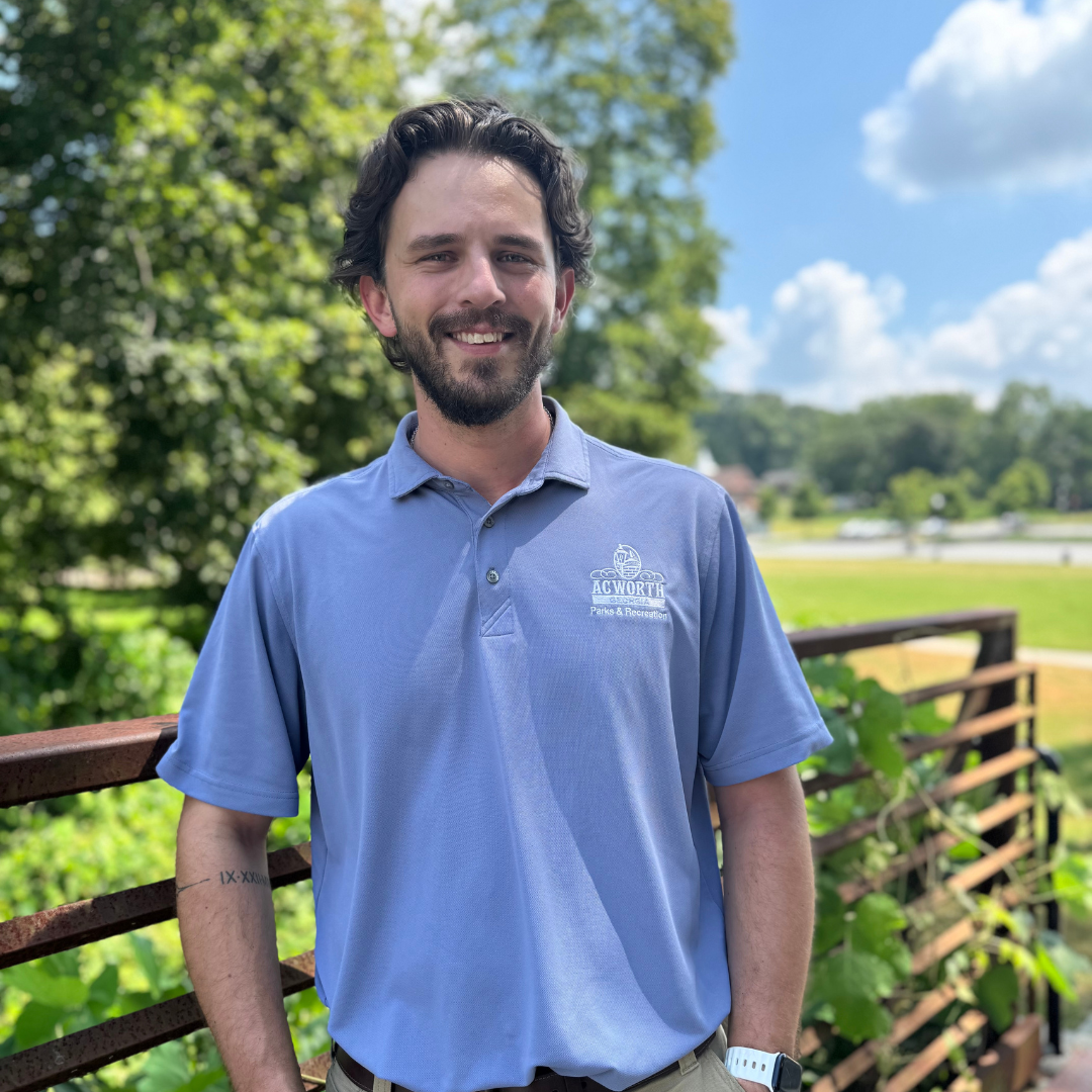 A man in a light blue polo shirt stands outdoors by a metal railing, with green trees and a grassy area in the background under a blue sky.