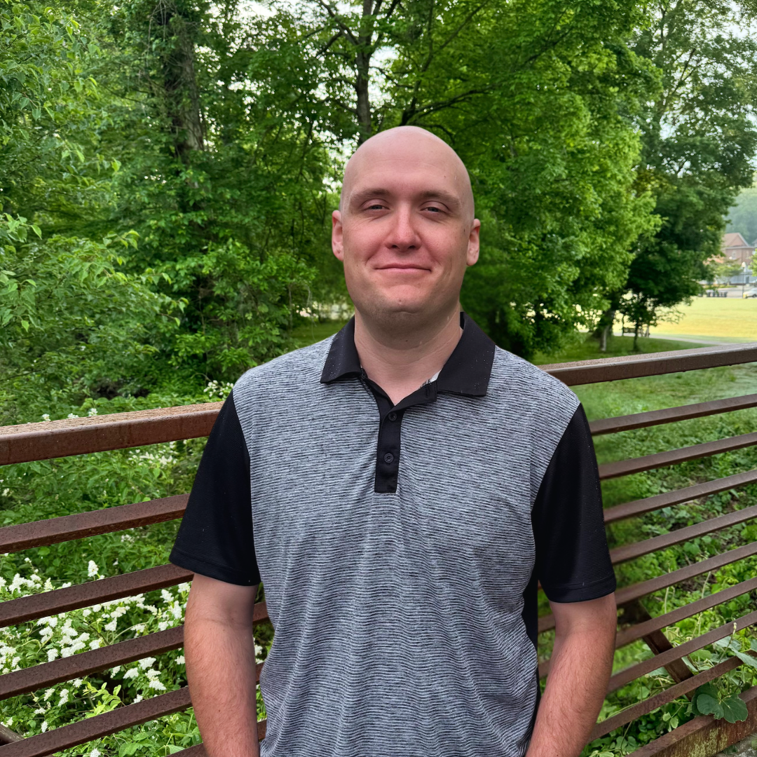 A man with a shaved head stands outside on a bridge, wearing a short-sleeve collared shirt, with trees and greenery in the background.