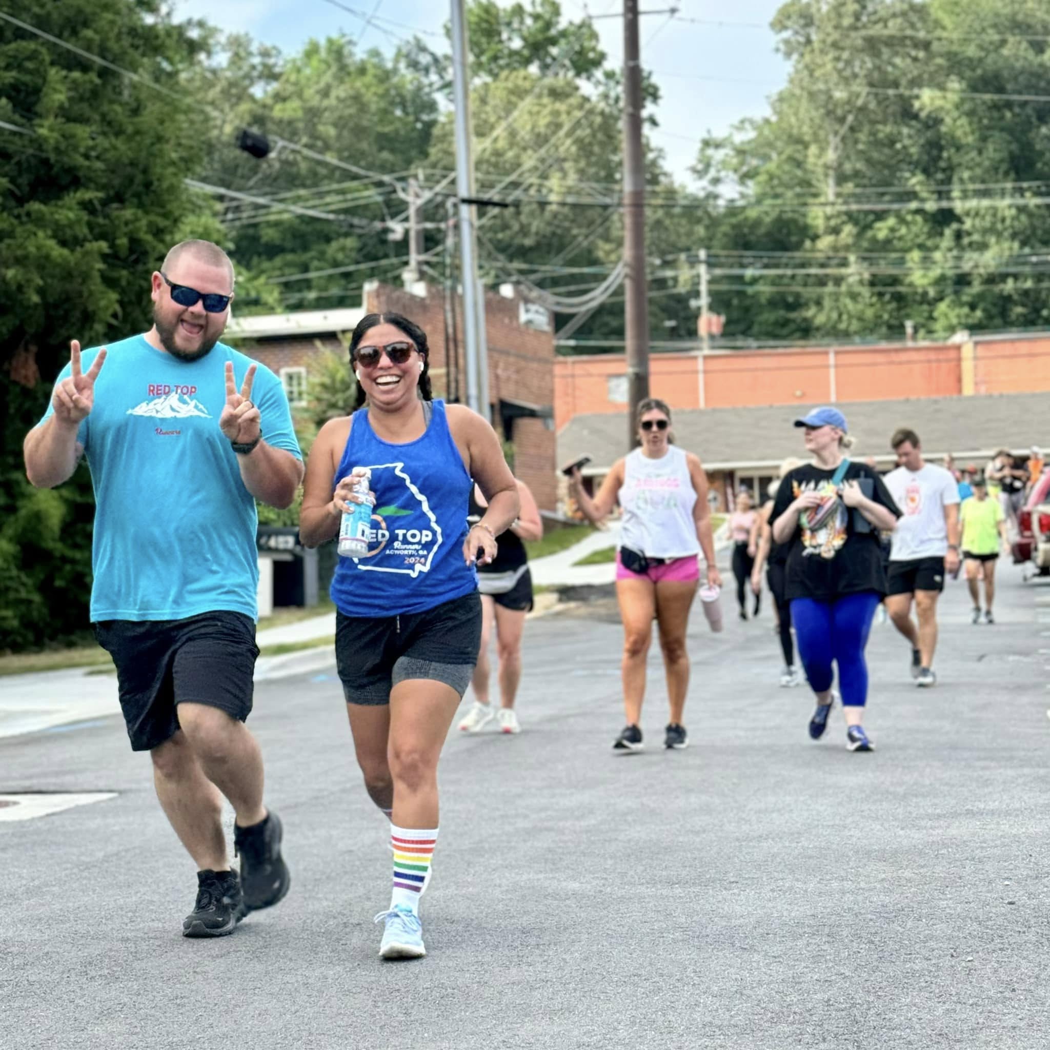 A group of people participates in an outdoor run or walk event; two people in front smile and make peace signs while others follow behind on a paved street.