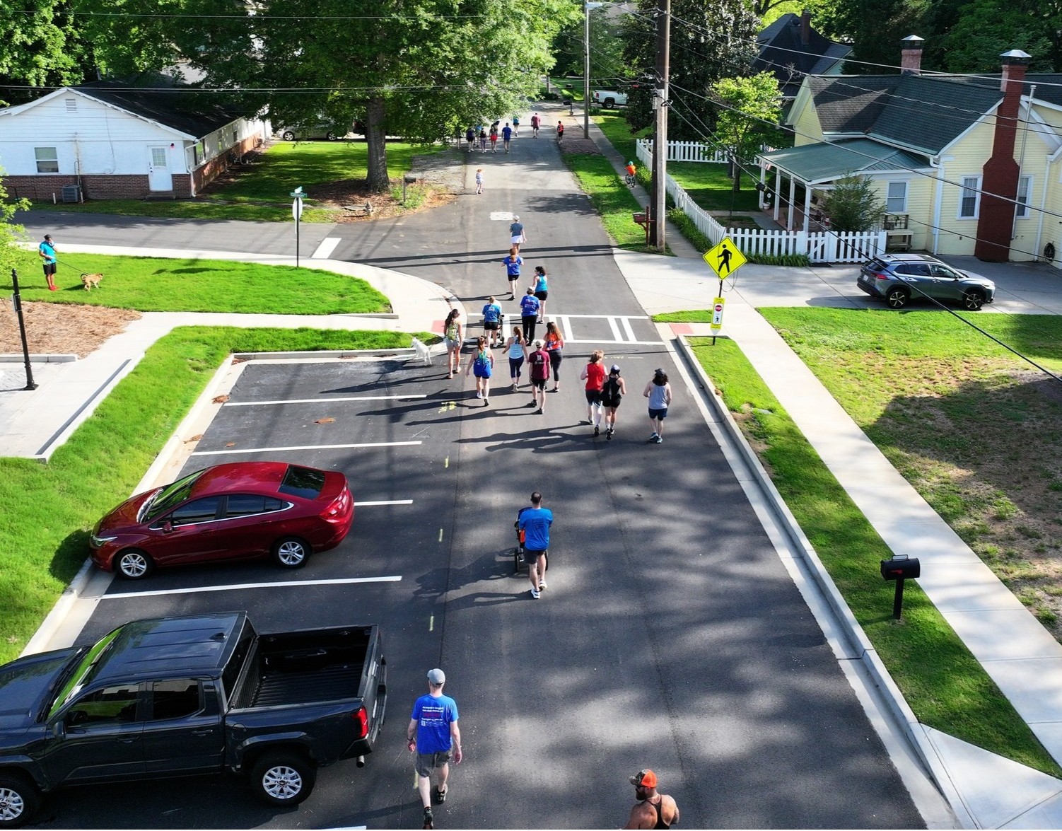 A group of people walk on a residential street closed to traffic, with parked cars on the side and houses lining the road.