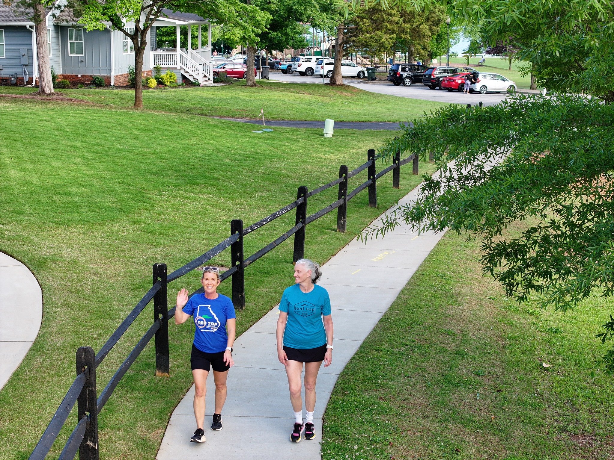 Two women walk on a curved sidewalk in a park-like area with grass, trees, parked cars, and houses in the background. Two women walk on a curved sidewalk in a park-like area with grass, trees, parked cars, and houses in the background.
