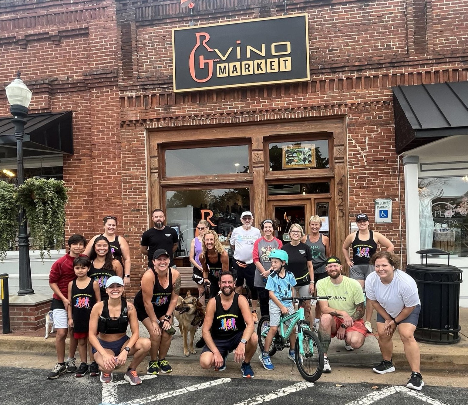 A group of people, some in athletic gear and one with a bicycle, pose for a photo outside a brick building labeled “G Vino Market.”.