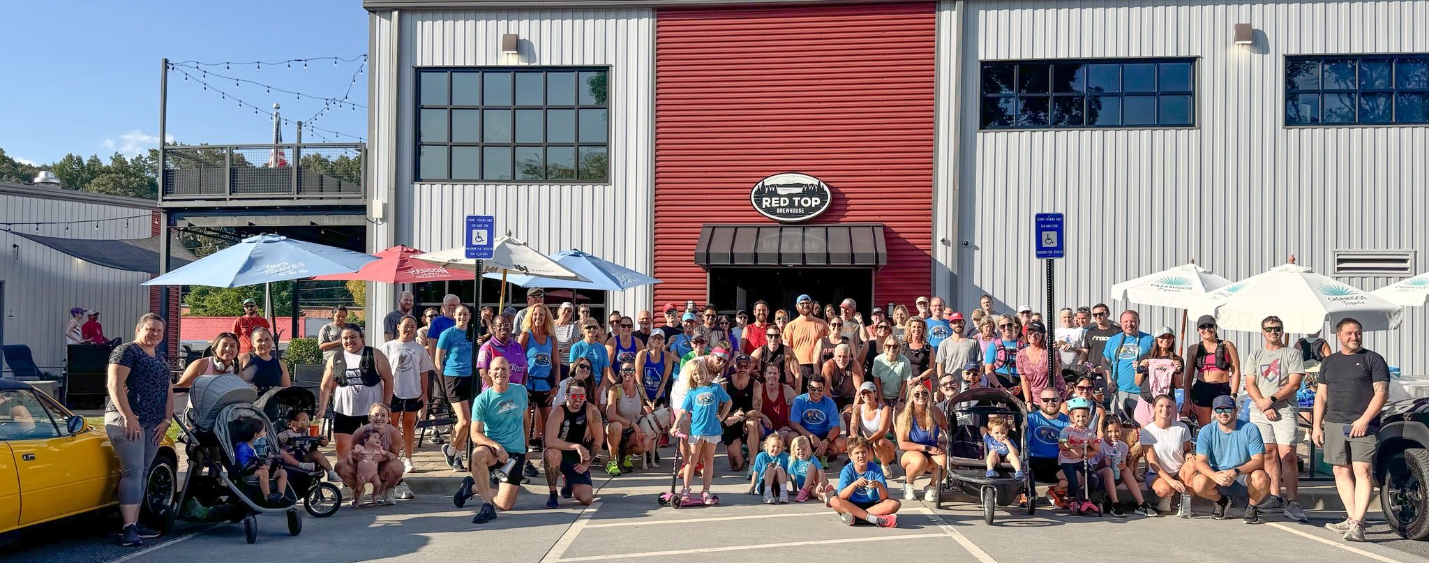 A large group of people, including adults and children, pose for a photo outside a red and gray building with "Red Top" signage on a sunny day.