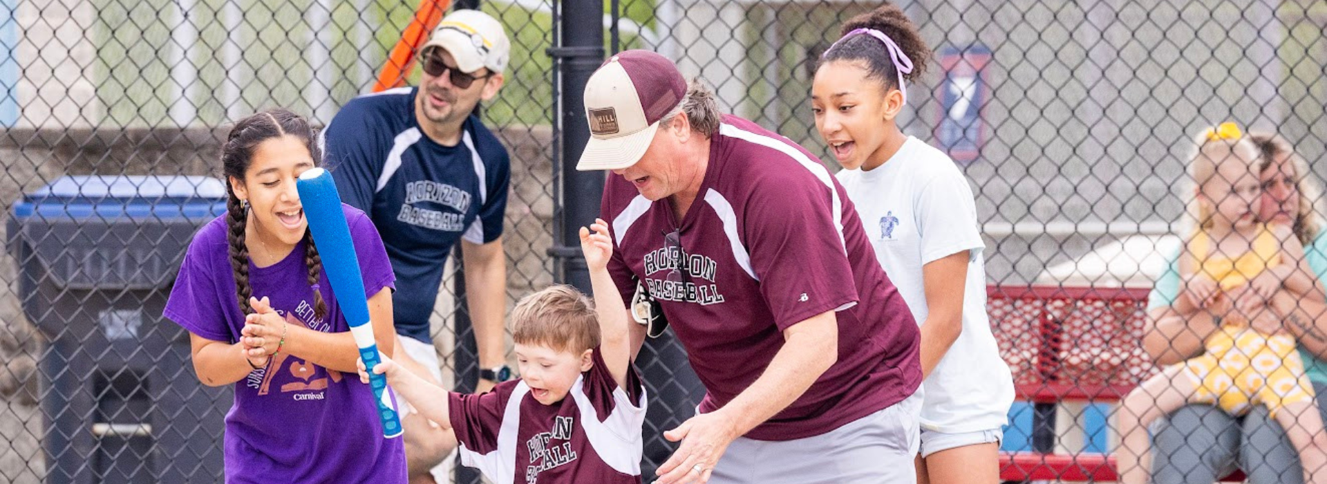 A young boy in a maroon sports jersey swings a bat with the guidance of an adult, while others cheer near a baseball field fence. A young boy in a maroon sports jersey swings a bat with the guidance of an adult, while others cheer near a baseball field fence.