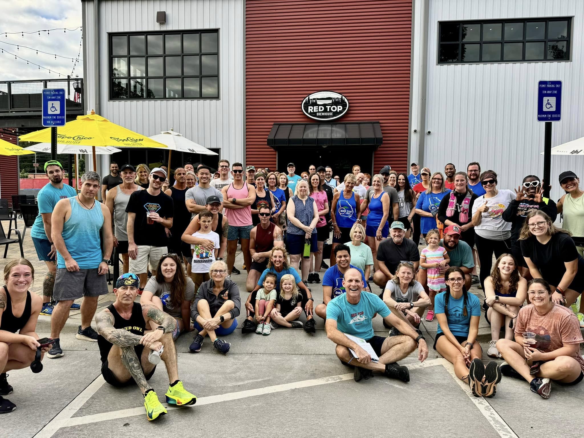 A large group of people pose and smile together outside a red and white building labeled “Red Top.” Some people are sitting or kneeling in front, while others stand behind them. A large group of people pose and smile together outside a red and white building labeled "Red Top." Some people are sitting or kneeling in front, while others stand behind them.