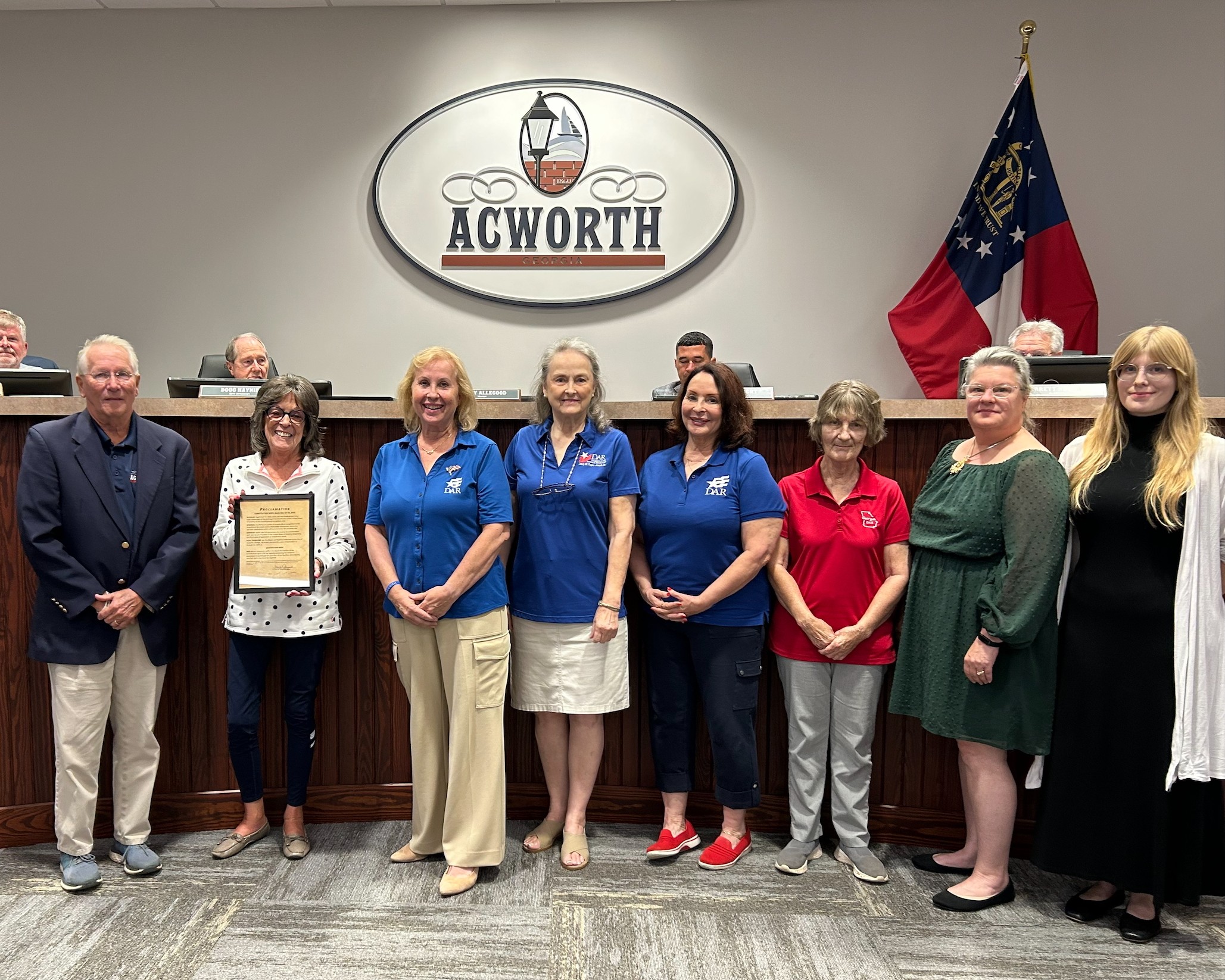 A group of people stands in front of a council dais and an Acworth sign; one person holds a framed document, with the Georgia state flag visible in the background.