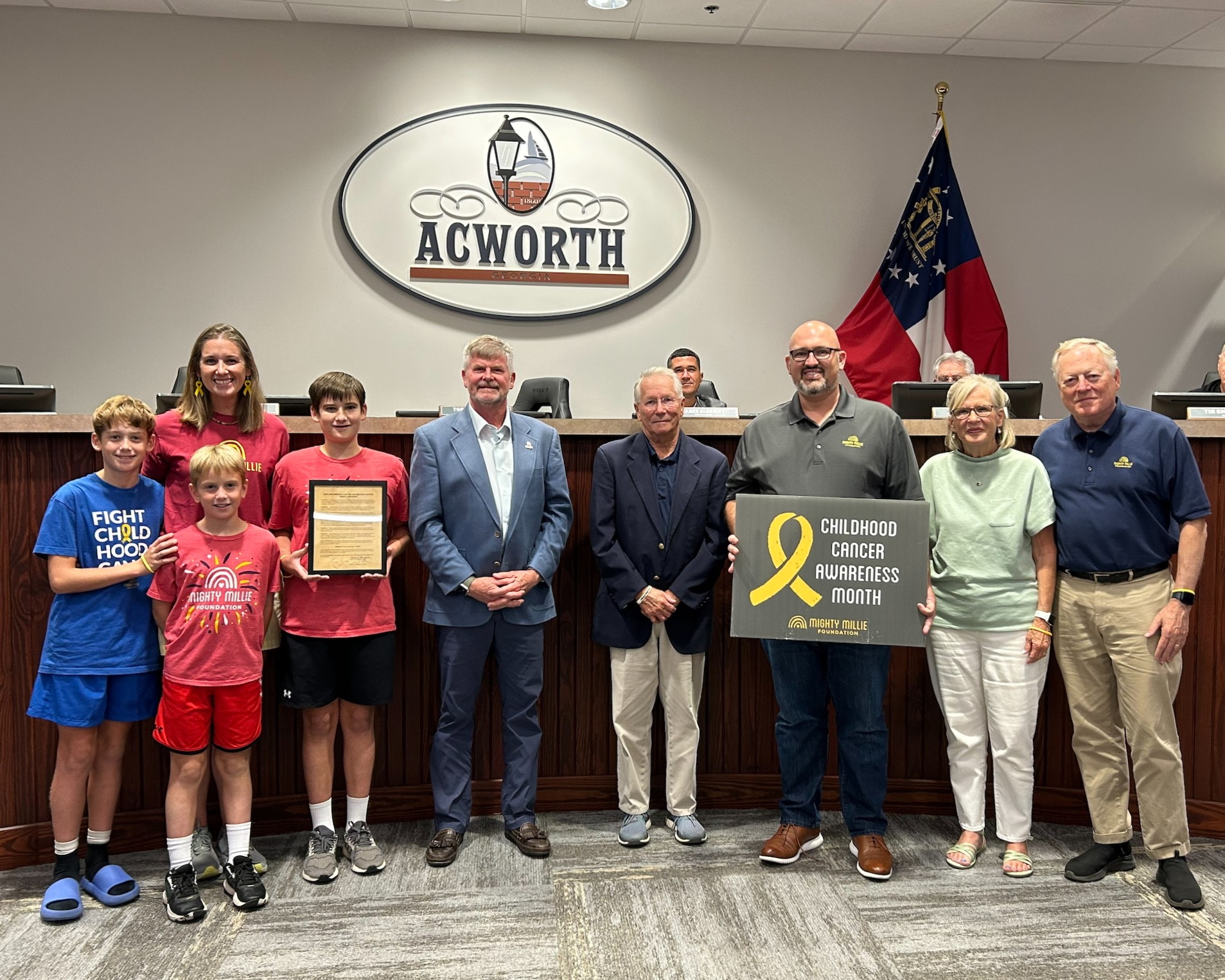 A group of people poses indoors, holding a plaque and a sign reading "Childhood Cancer Awareness Month," in front of a city council bench with an "Acworth" sign and Georgia flags.