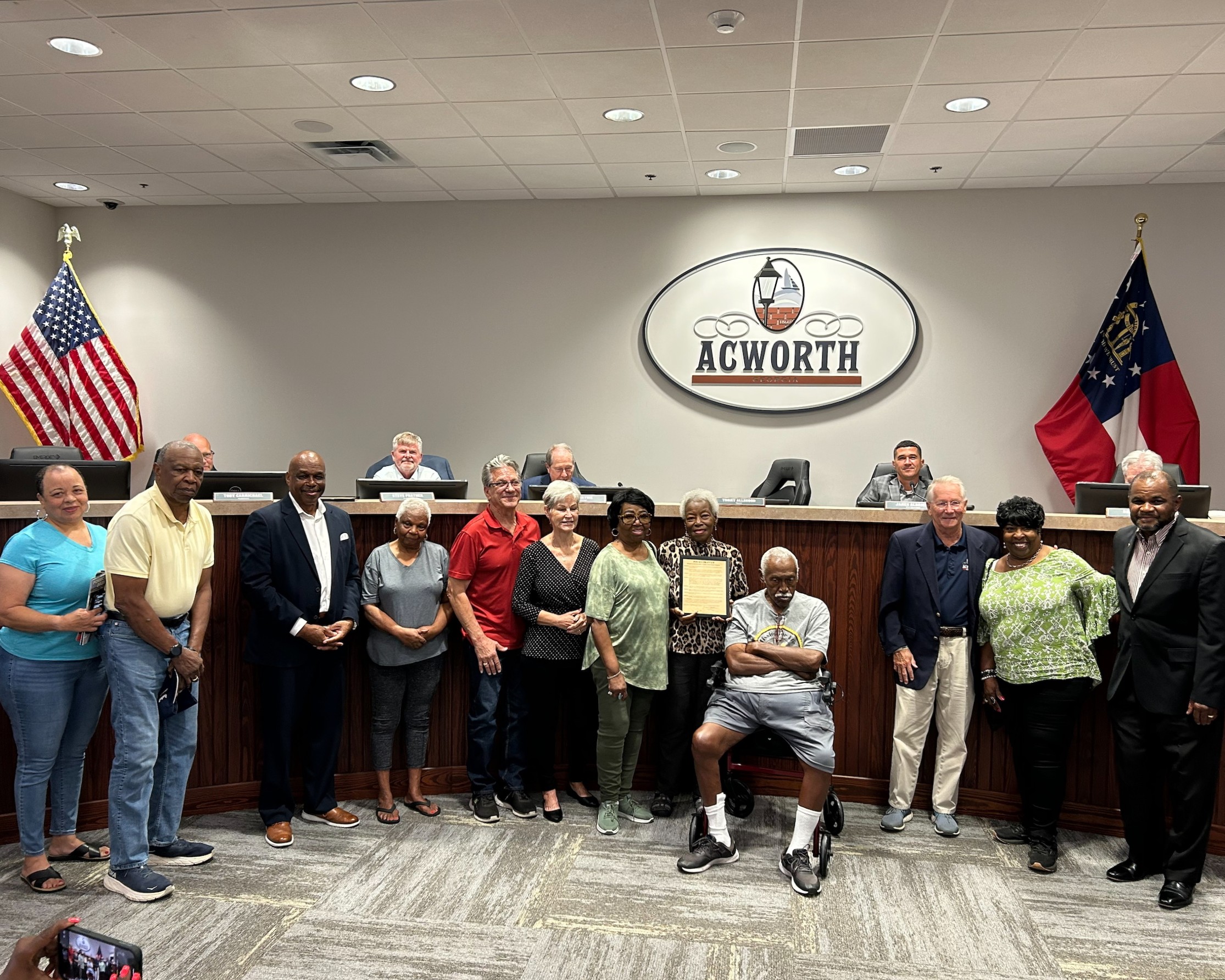 A group of people poses for a photo in a government chamber in front of an "Acworth" sign, with two flags and several officials seated behind them. One person holds a framed certificate.