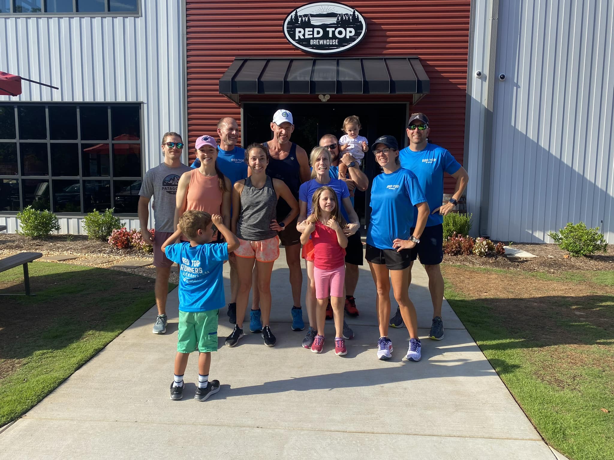 A group of people, including adults and children, stand and pose for a photo on a sidewalk outside a building with a “Red Top” sign. A group of people, including adults and children, stand and pose for a photo on a sidewalk outside a building with a "Red Top" sign.