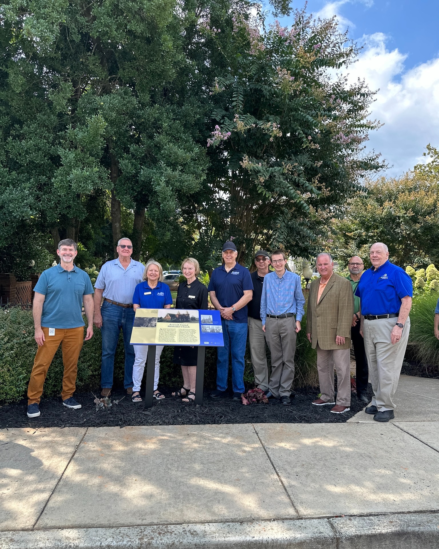 A group of eleven people stands outdoors on a sidewalk in front of trees and shrubs, gathered around an informational sign.