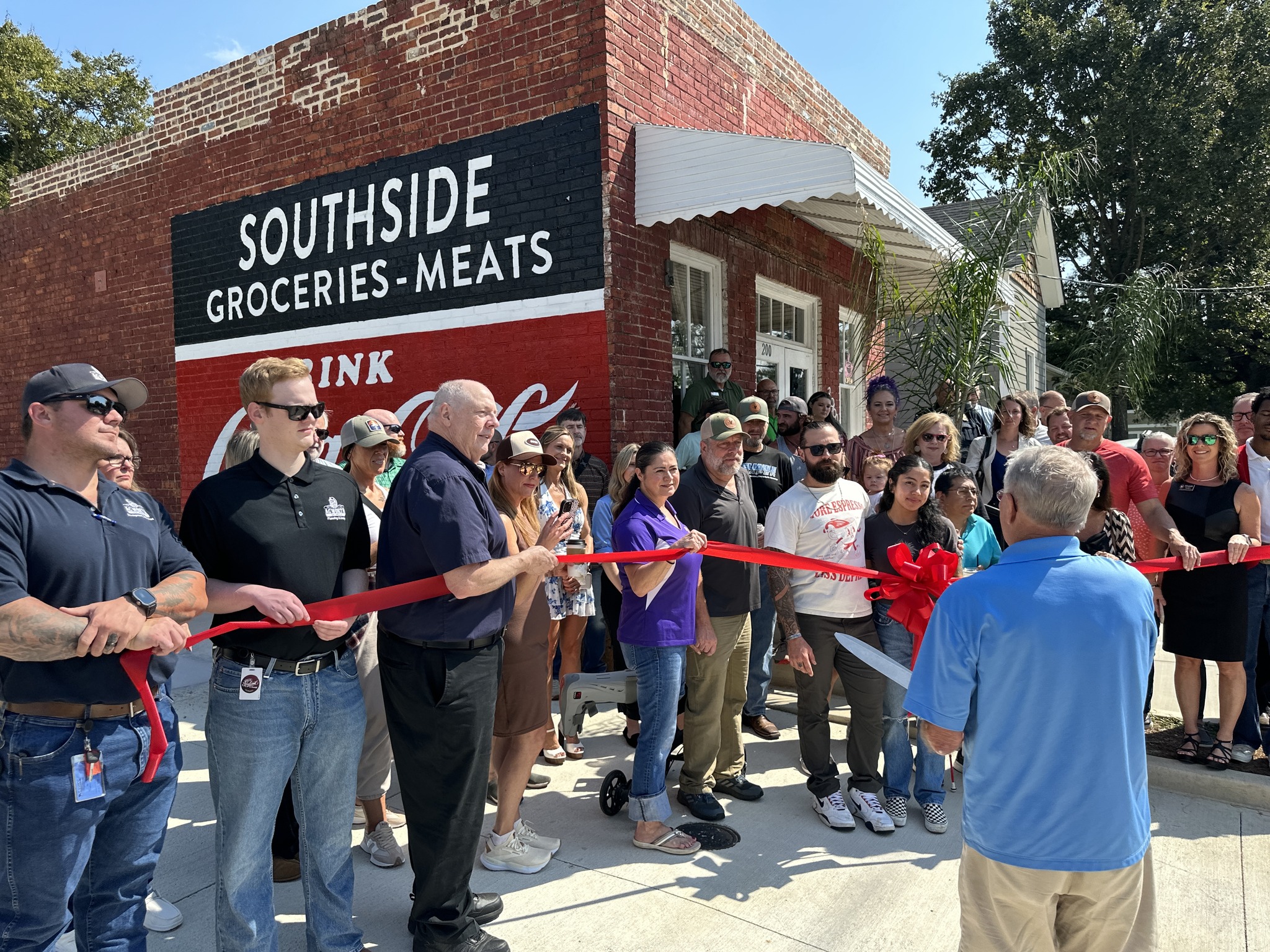 A group of people gathers outside Southside Groceries-Meats for a ribbon-cutting ceremony, with one man holding large scissors.