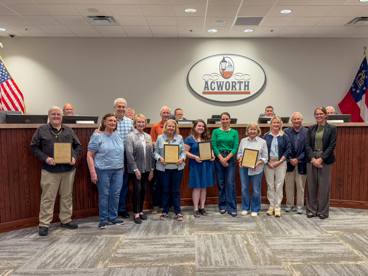 A group of people pose indoors at the Acworth city council chamber, with three individuals in front holding plaques, and a city seal and flags visible in the background.