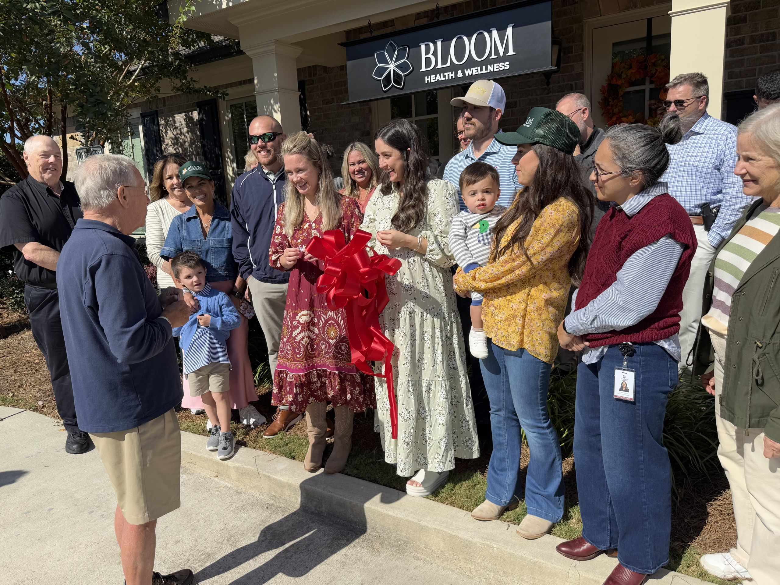 A group of people gathers outside Bloom Health & Wellness for a ribbon-cutting ceremony. A woman holds large scissors, preparing to cut a red ribbon.