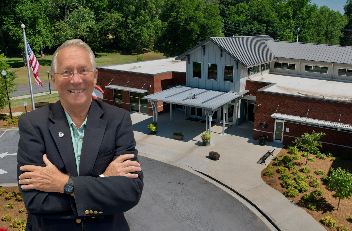 Smiling man in a blazer stands with arms crossed in front of a modern brick building with a metal roof and flagpole.