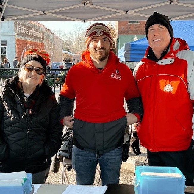 Three people wearing jackets stand behind a table with paperwork and bins at an outdoor event. A tent, fence, and other people are visible in the background. Three people wearing jackets stand behind a table with paperwork and bins at an outdoor event. A tent, fence, and other people are visible in the background.