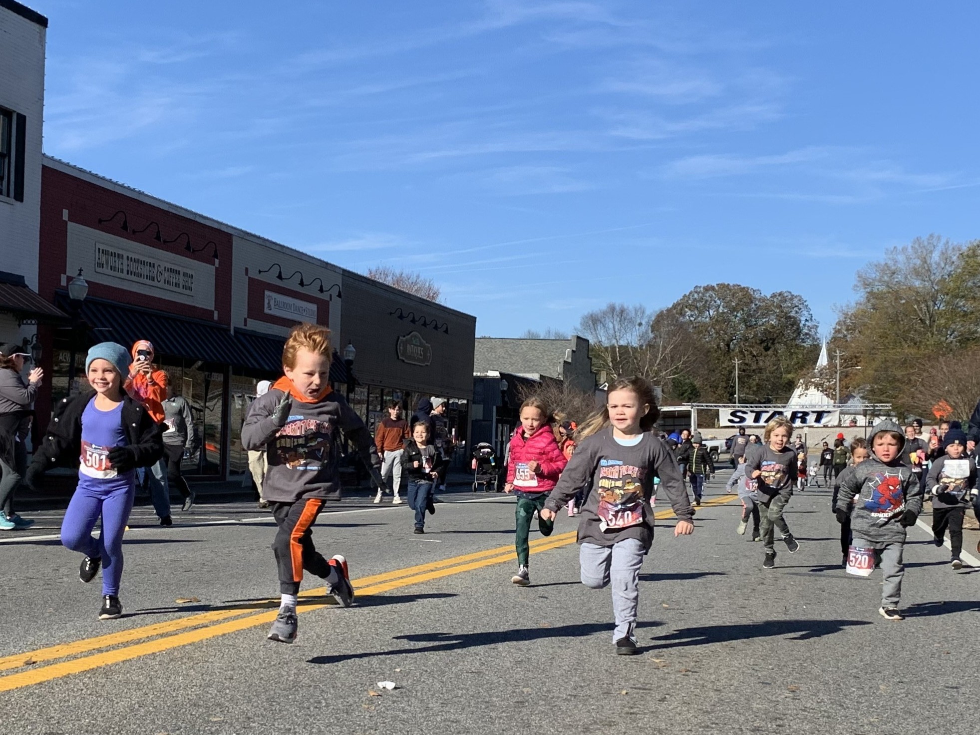 Children run a race on a street lined with buildings, with spectators watching and a “START” banner visible in the background. Children run a race on a street lined with buildings, with spectators watching and a "START" banner visible in the background.