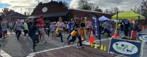 A group of runners, some in costumes, cross the starting line at an outdoor race event on a paved street near a brick building.