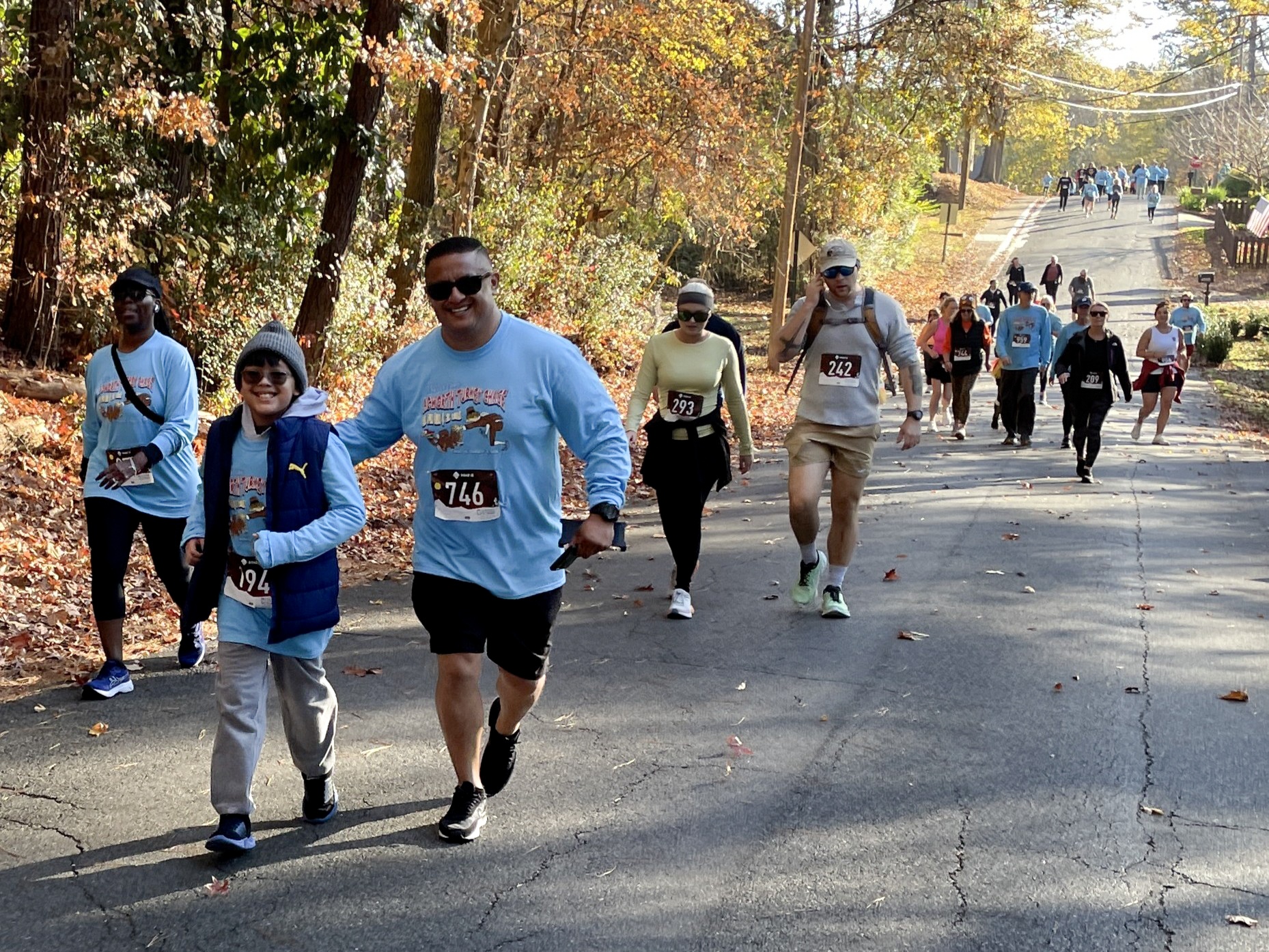 A group of people, some wearing race bibs, walk up a tree-lined street during a fall outdoor event. A group of people, some wearing race bibs, walk up a tree-lined street during a fall outdoor event.