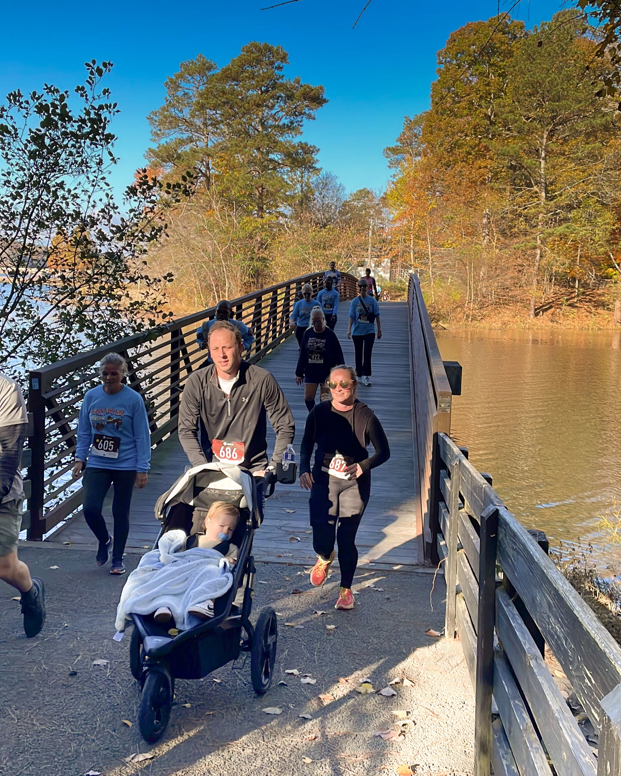 Runners cross a wooden bridge over a river during a race; one man pushes a stroller with a child, and other participants follow behind on a sunny day. Runners cross a wooden bridge over a river during a race; one man pushes a stroller with a child, and other participants follow behind on a sunny day.