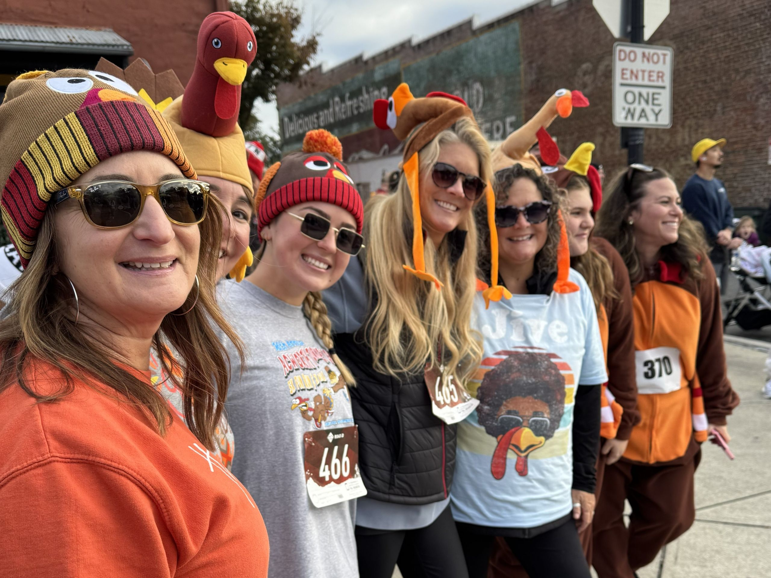 A group of women in turkey-themed hats and shirts pose and smile outdoors at a festive event, with race bibs visible on their shirts. A group of women in turkey-themed hats and shirts pose and smile outdoors at a festive event, with race bibs visible on their shirts.