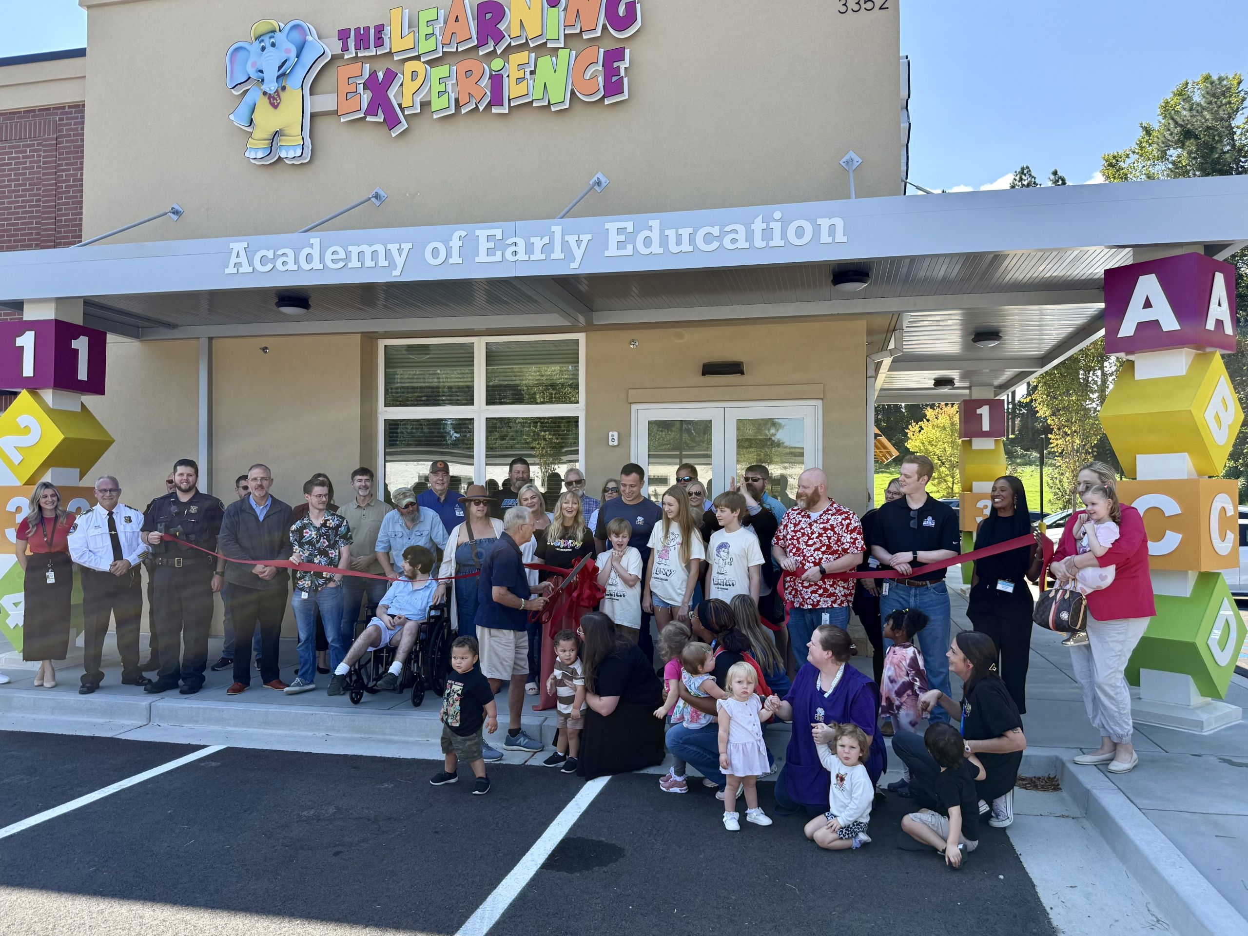 A group of adults and children gather for a ribbon-cutting ceremony outside The Learning Experience Academy of Early Education building.