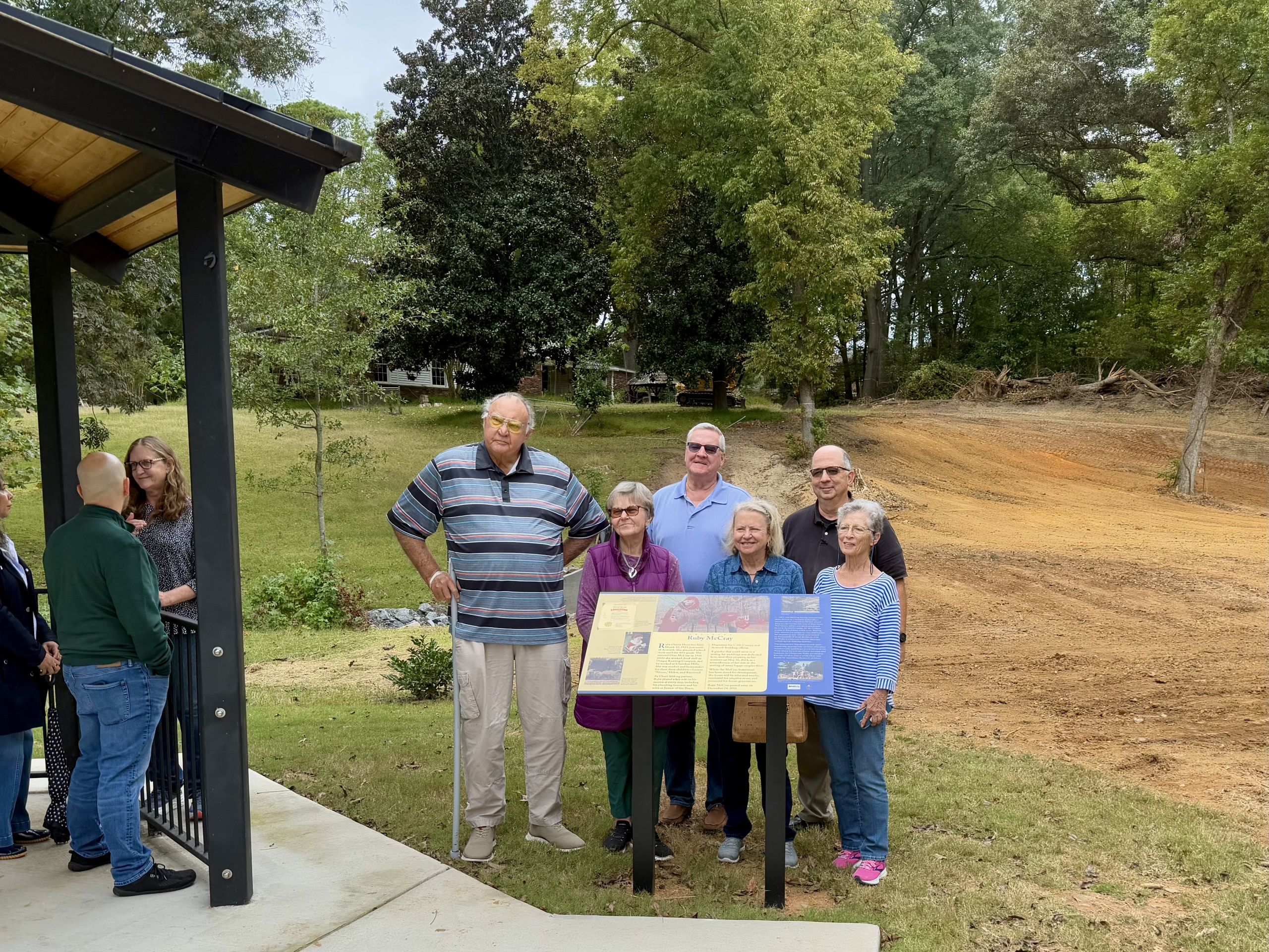 A group of seven adults stand next to informational signs at an outdoor park area with trees and an open dirt field in the background.