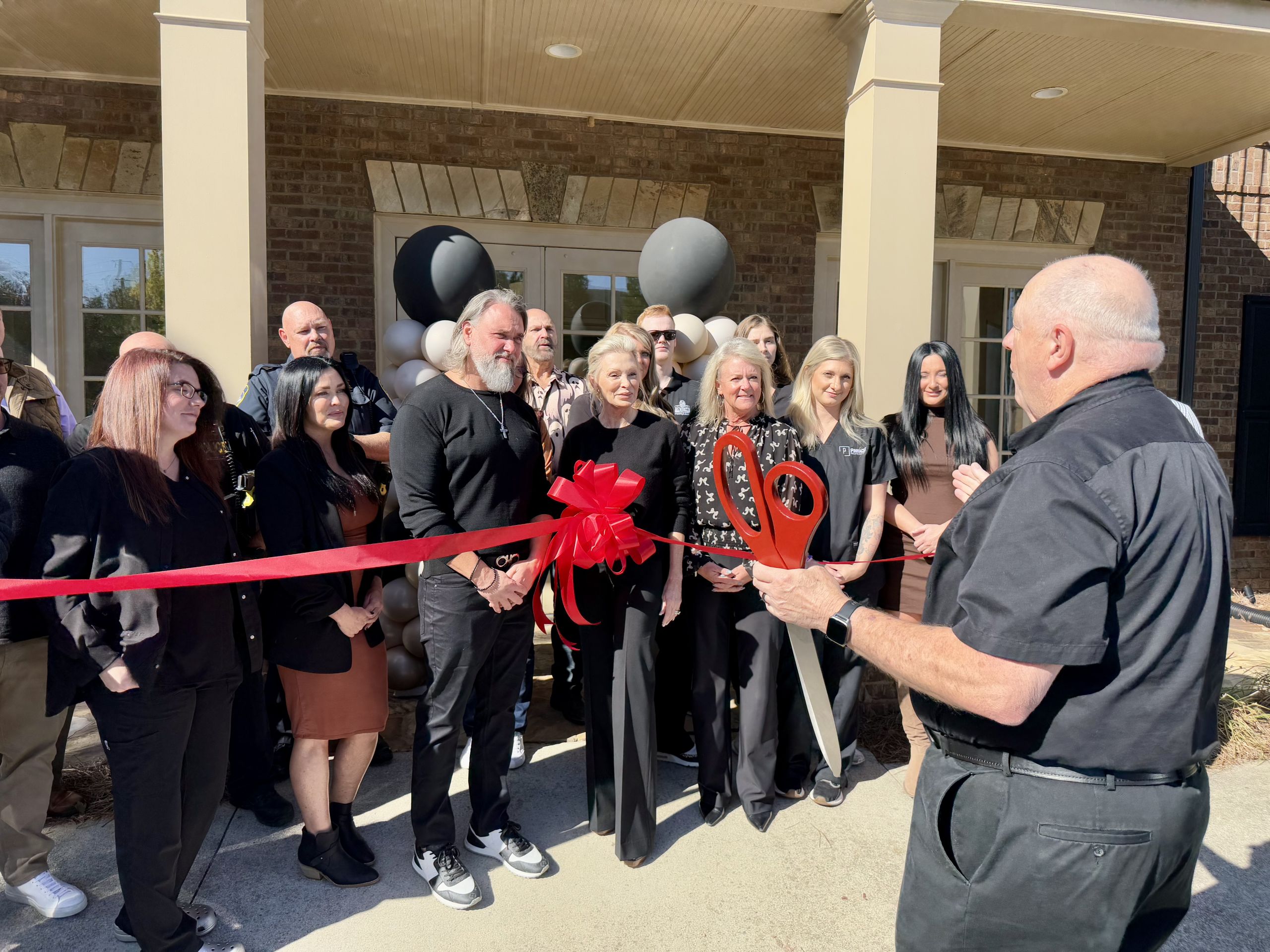 A group of people stands outside a building as a man prepares to cut a red ribbon with large scissors during a ribbon-cutting ceremony.