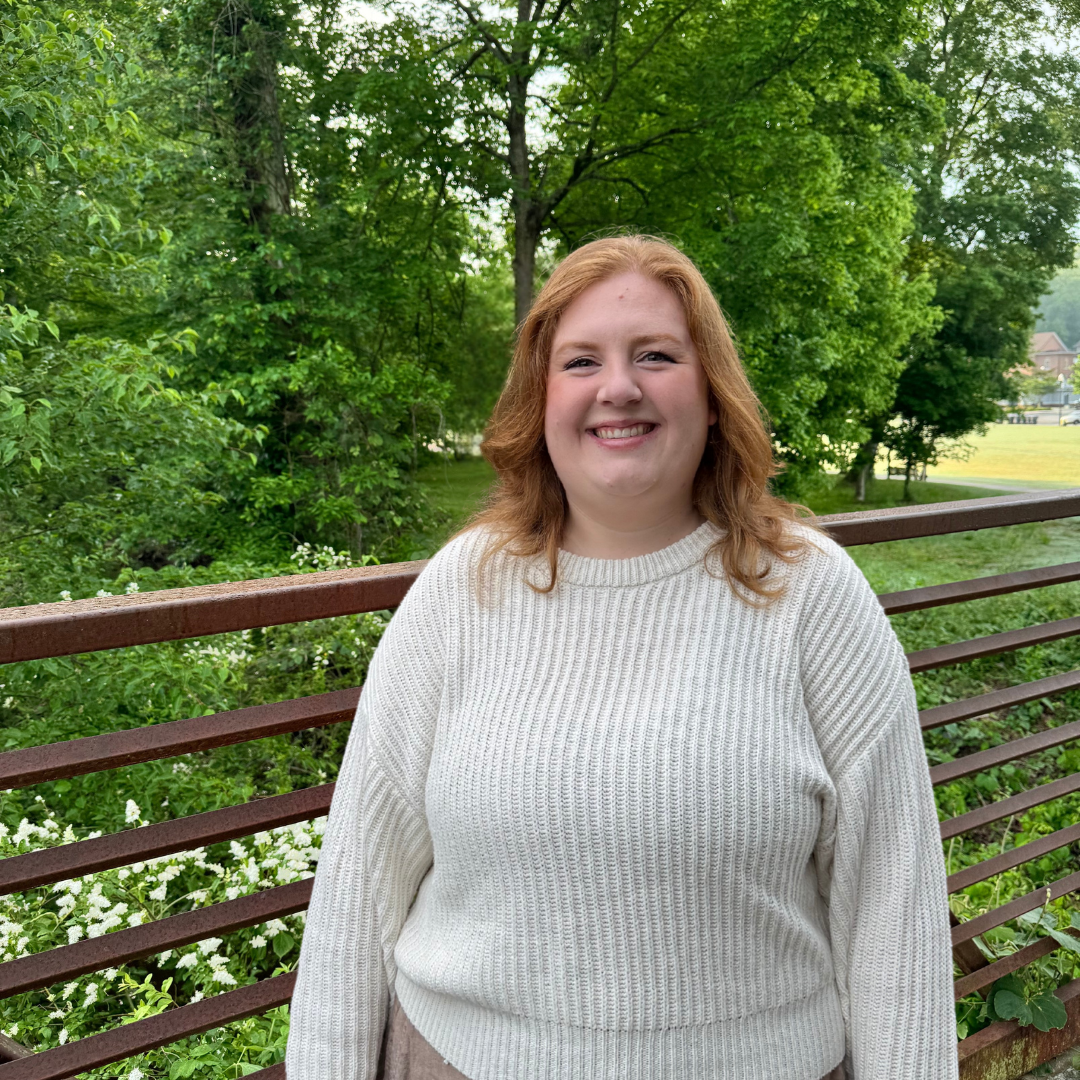 A person with shoulder-length red hair, wearing a white sweater, stands outdoors in front of a metal railing with green trees in the background.