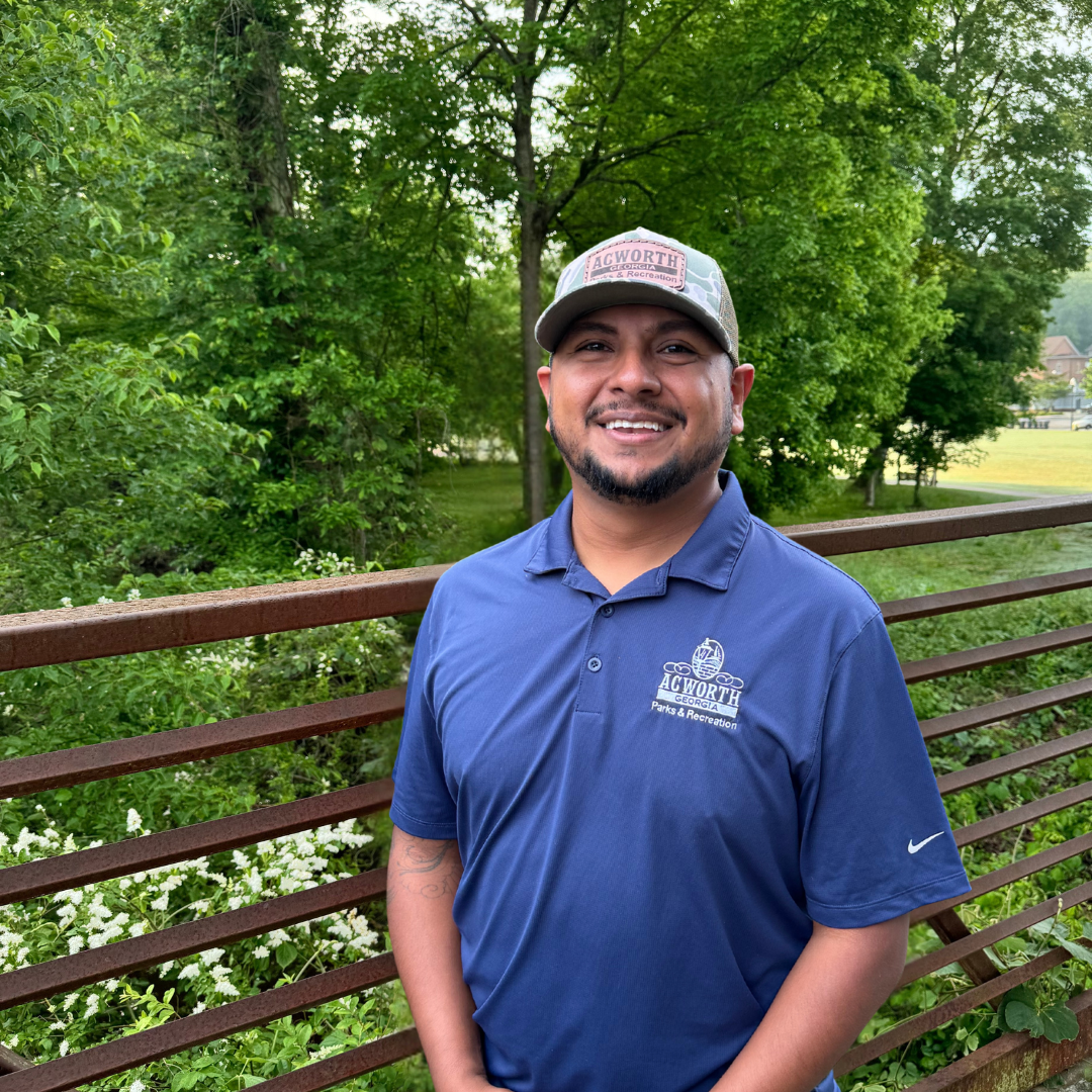 A man in a blue polo and cap stands smiling on a bridge with green trees and grass in the background.