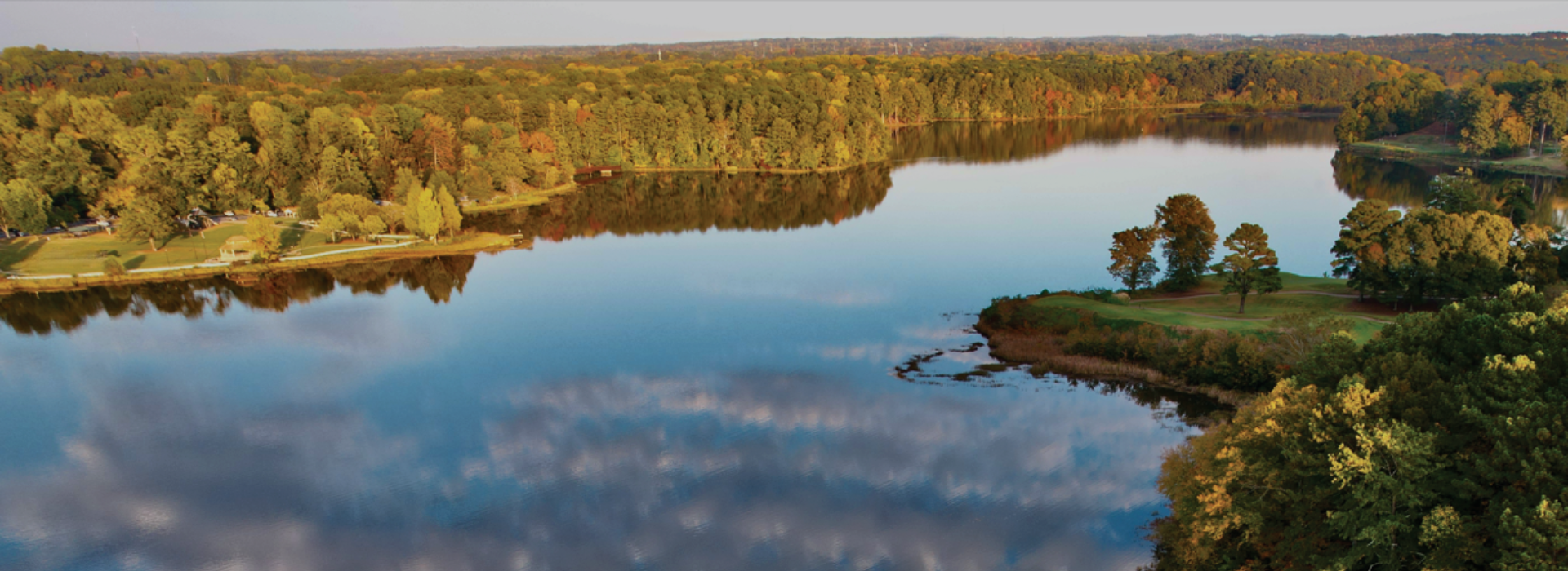 A calm lake reflects the sky and surrounding trees, bordered by dense forest and a grassy area, under a clear sky. A calm lake reflects the sky and surrounding trees, bordered by dense forest and a grassy area, under a clear sky.