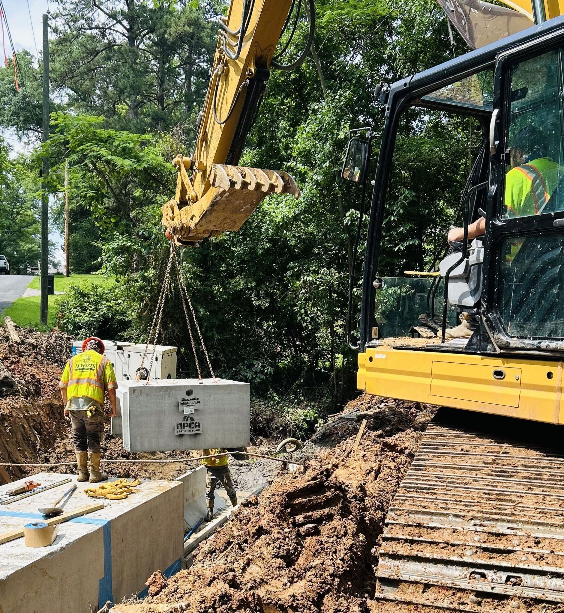 A construction worker operates an excavator to lower a concrete box into a trench while two other workers guide it into place. A construction worker operates an excavator to lower a concrete box into a trench while two other workers guide it into place.