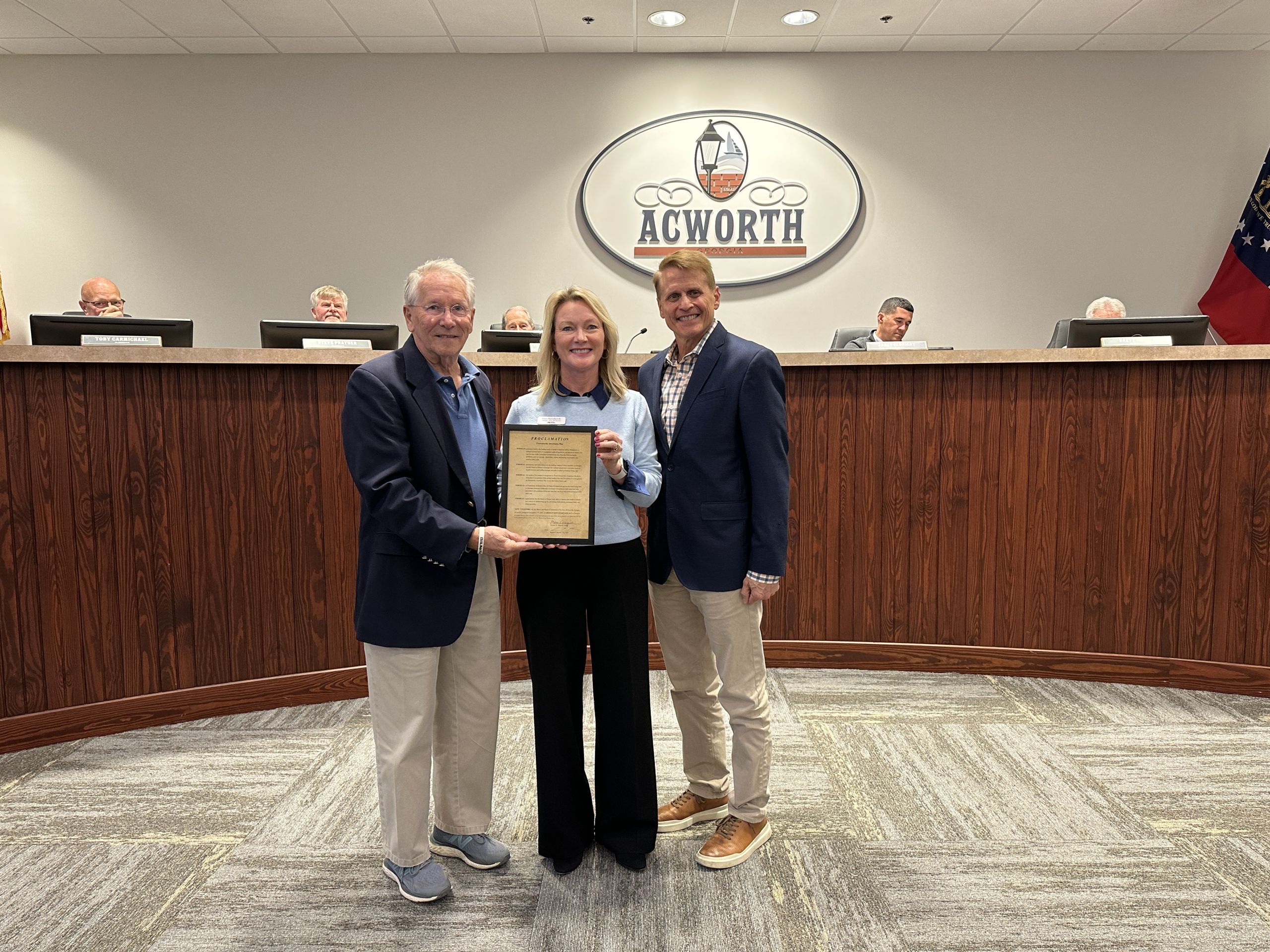 Three people pose holding a framed document in front of a city council bench with an "Acworth" sign on the wall behind them.