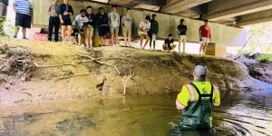 A person in waders stands in a shallow creek under a bridge, speaking to a group of people gathered on a dirt embankment.