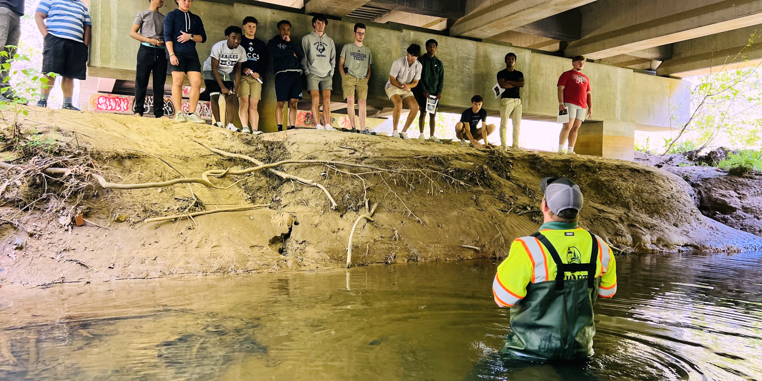A person in waders stands in a shallow creek under a bridge, speaking to a group of people gathered on a dirt embankment. A person in waders stands in a shallow creek under a bridge, speaking to a group of people gathered on a dirt embankment.