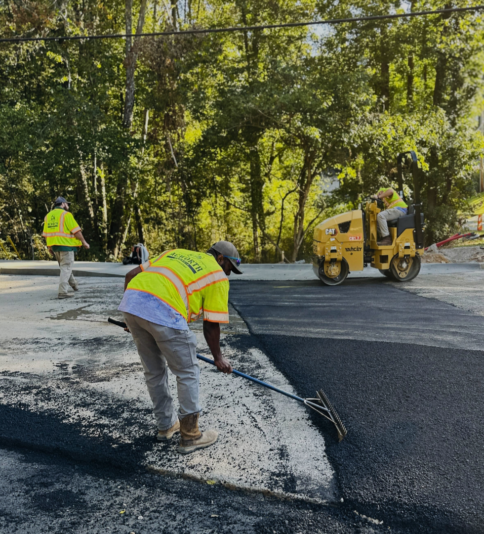 Three construction workers repave a road; one rakes asphalt while another operates a steamroller, with trees and equipment visible in the background. Three construction workers repave a road; one rakes asphalt while another operates a steamroller, with trees and equipment visible in the background.