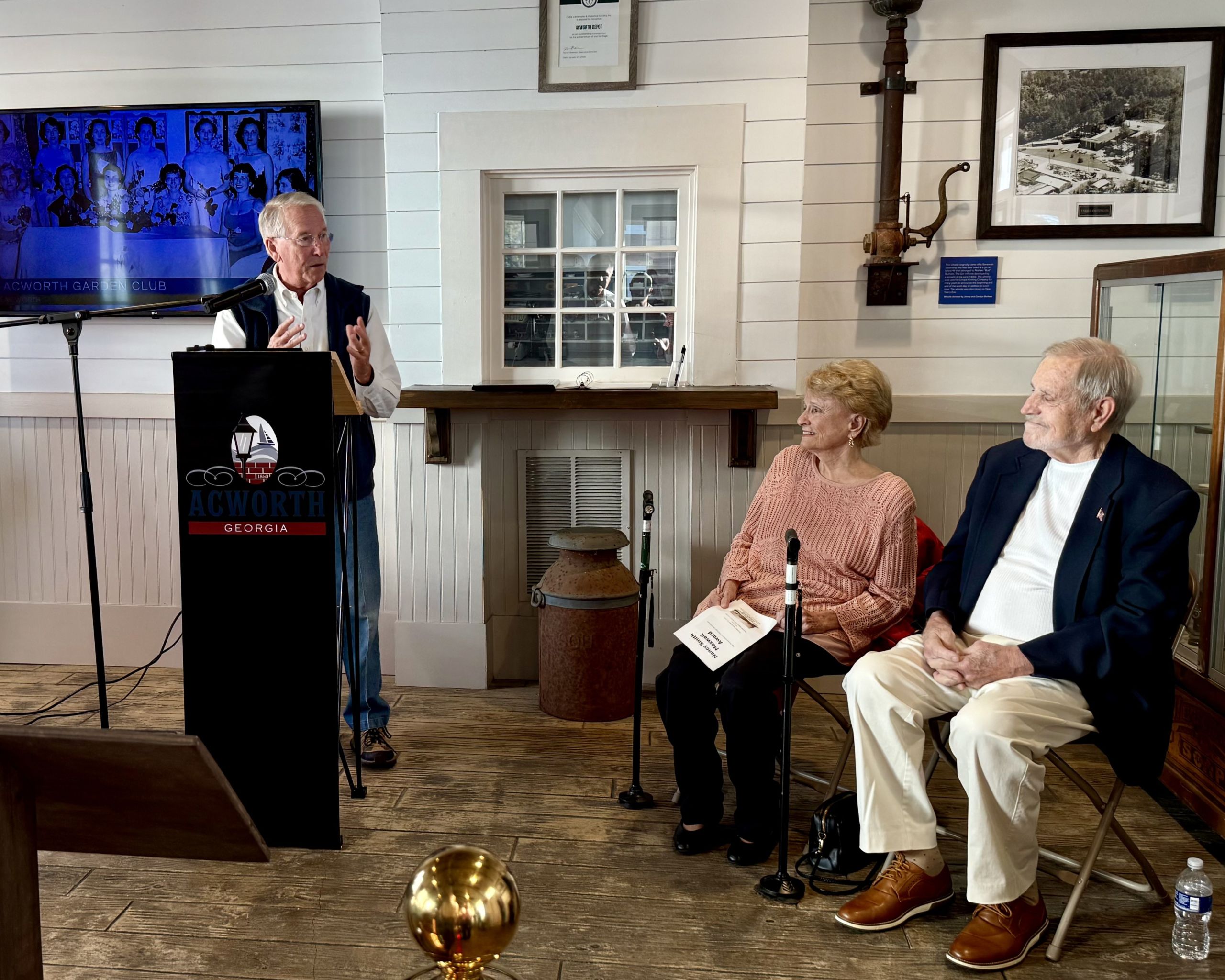 An older man speaks at a podium labeled "Acworth Georgia", while an elderly woman and man sit and listen in a room decorated with historical photos and artifacts.