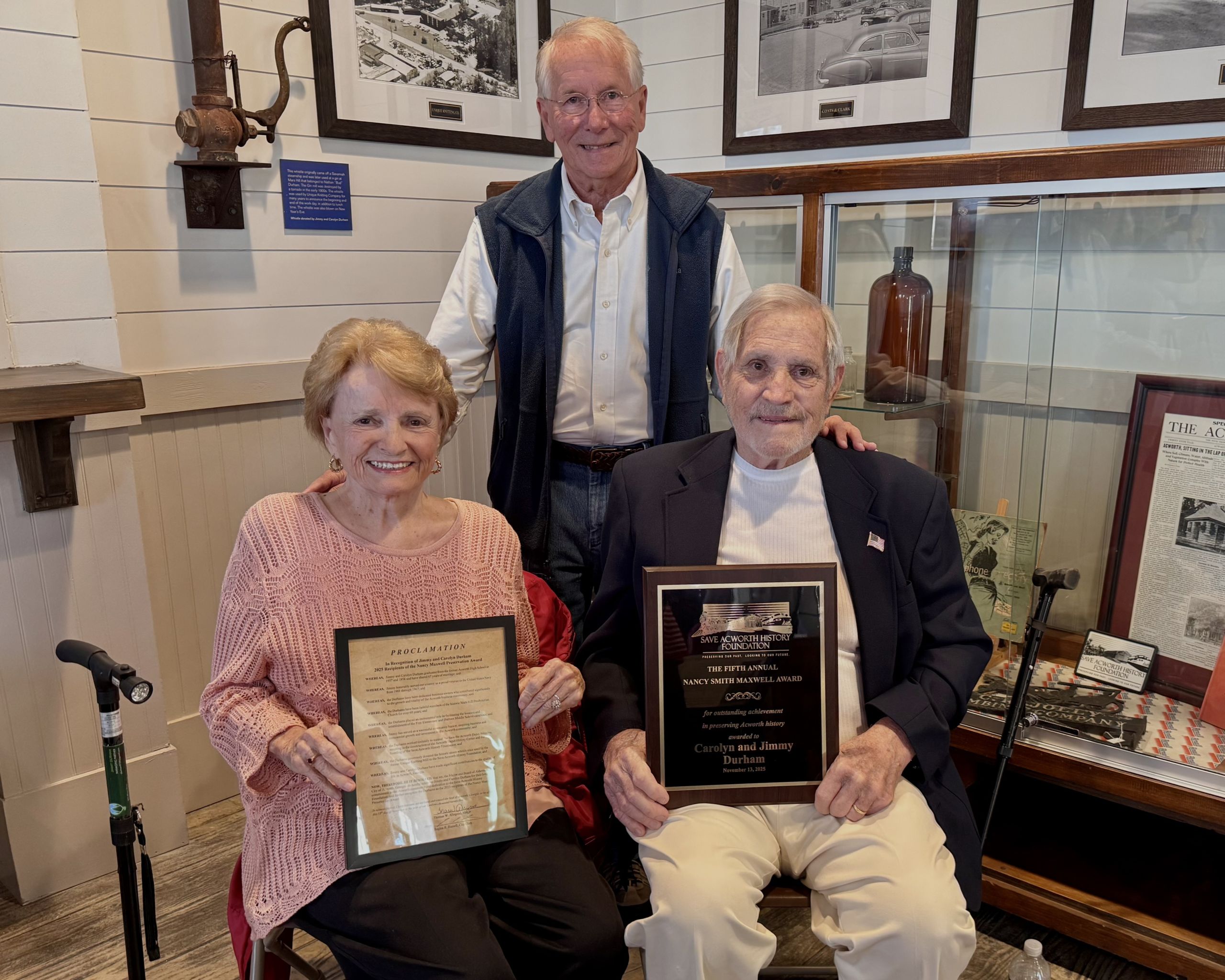Three older adults pose indoors; the woman on the left holds a framed certificate, the man on the right holds a plaque, and another man stands behind them. Historic photos are visible in the background.